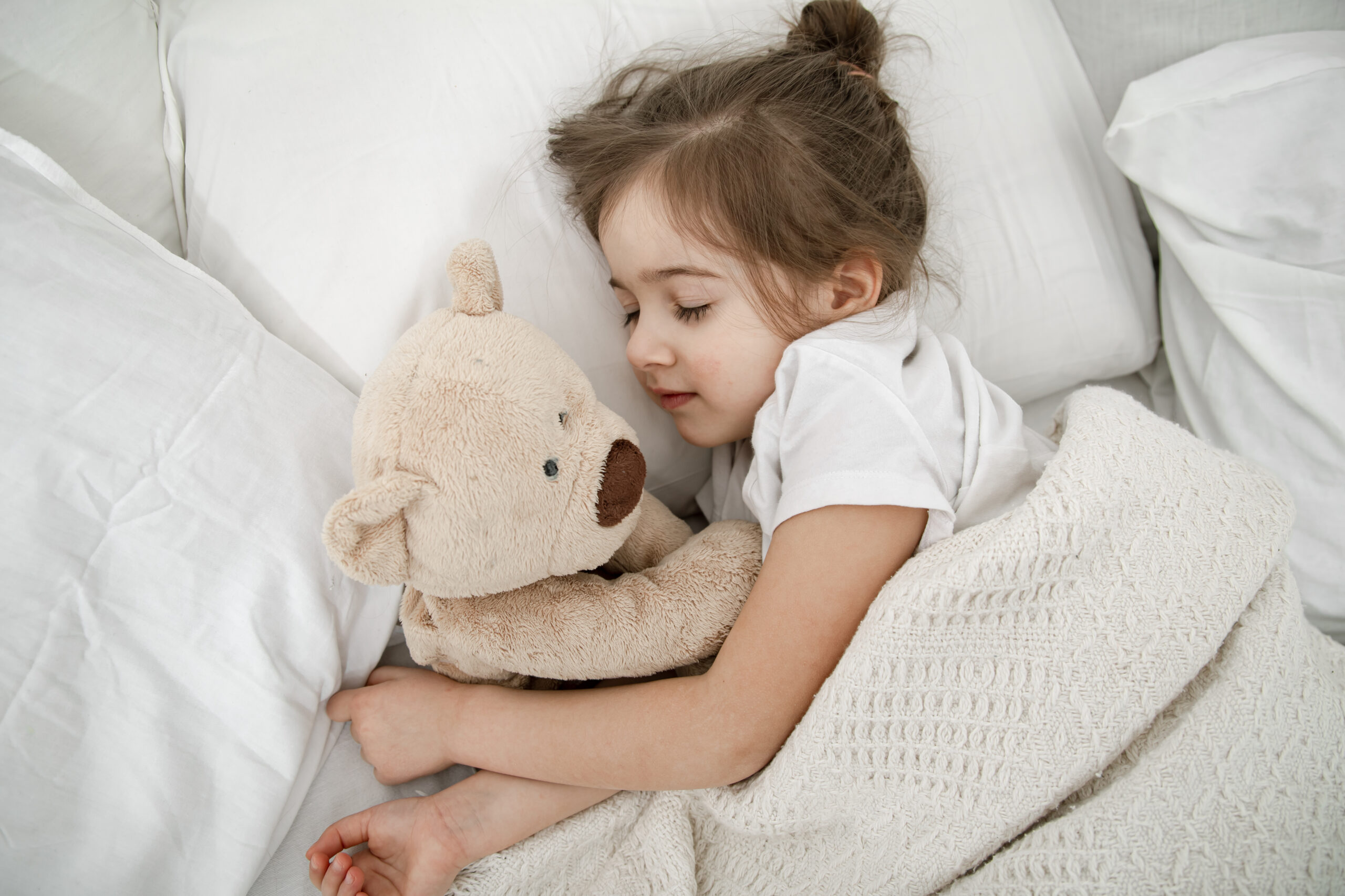 A cute little girl is sleeping in a bed with a Teddy bear toy . Concept of child development and sleep. The view from the top.