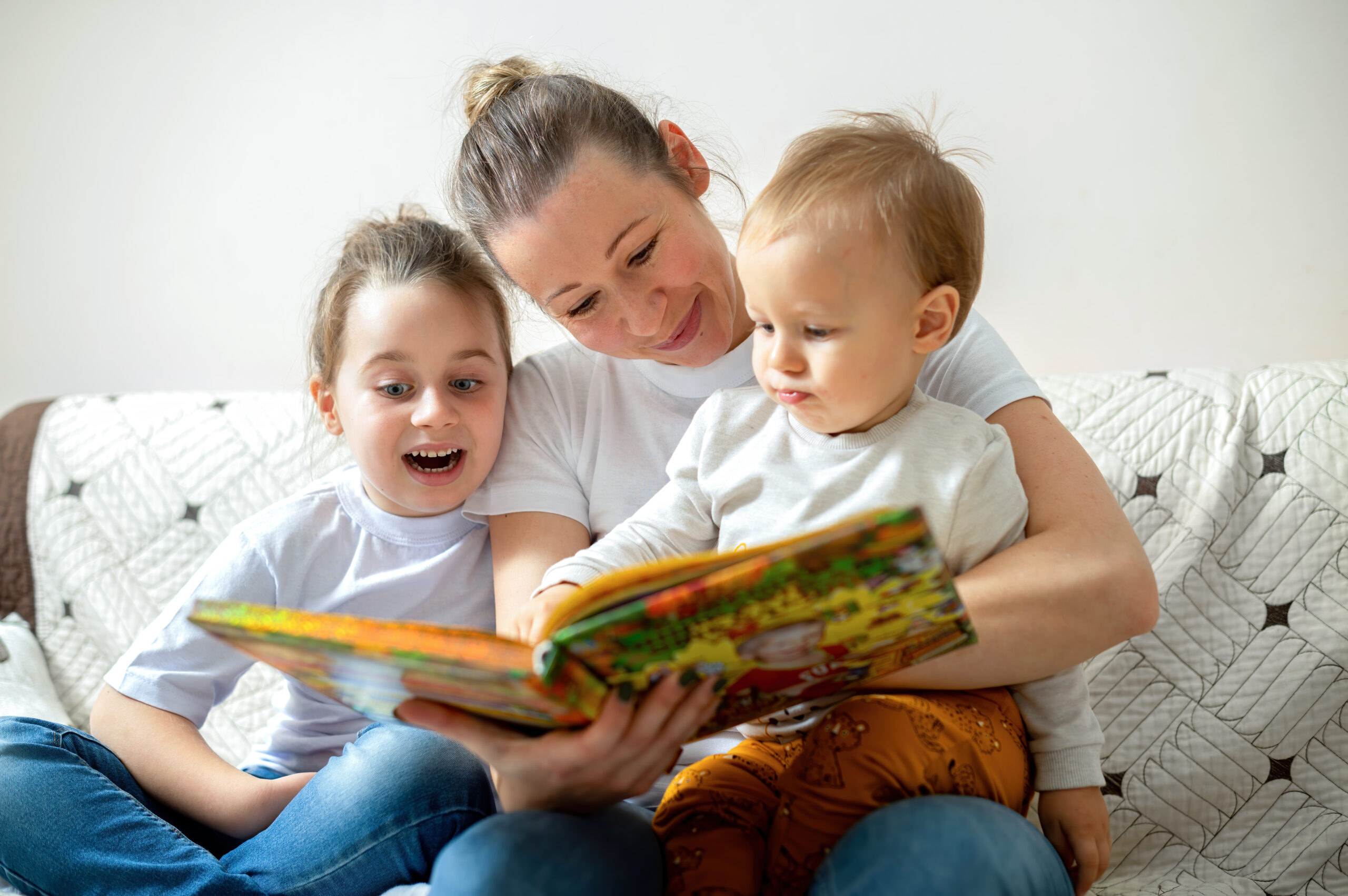 Mom and her two children are reading a book at home on a sofa. Happy and smiling