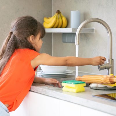 Cute little girl washing dish in kitchen by herself. Child reaching kitchen sink faucet tap and turning on water. Domestic shores concept